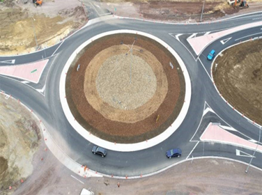 Bird's eye view of a newly constructed roundabout with a big dirt patch in the center. 3 cars are going around it