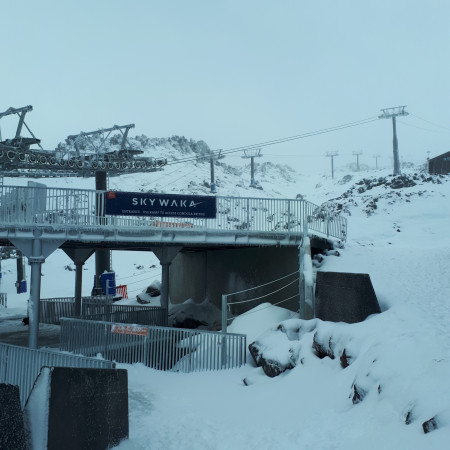 An entryway with metal barriers, leading to a gondola, among thick snow and rocks, on top of a mountain. The entryway has a dark blue banner that says 'Sky Waka