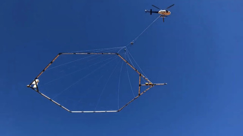 Low angle photo of a helicopter with a hexagonal object hanging from it. Vibrant blue sky above