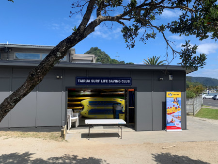Modern, grey building filled with surf lifesaving paddle boards and a sign saying 'Tairua Surf lifesaving Club' with a tressel table and a tree in front of it.