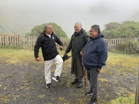 Three men in wet weather gear stand on a patch of grass and gravel with a fence, trees and rainy mist in the background