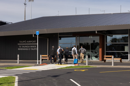 Image of the outside of the Taupō Airport - a small, modern building with a new road outside and a group of people walking in the door, carrying luggage.