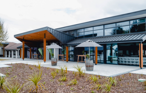 Image of a modern building with wine barrels holding umbrellas outside, and freshly planted plants in front of the building.