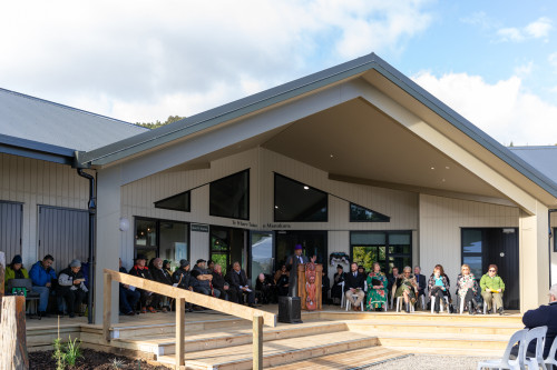 A man wearing a suit and a purple hat speaks at a lecturn with a Māori carving on the front of it. He stands in front of a large group of people sitting on chairs on the newly built deck of the new building.