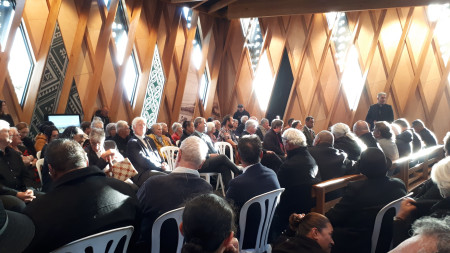 Large group of people in formal clothing sitting in a room with wooden walls and Māori designs on the walls. They are listening to one person who is standing and talking.