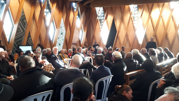 A large group of people sitting on chairs inside a building with wooden diamond panelling and Māori and Pasifika designs.