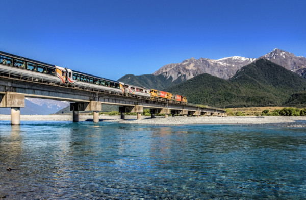 Low angle shot of a KiwiRail train going across a bridge with clear blue shallow water underneath, and snowy mountains in the distance